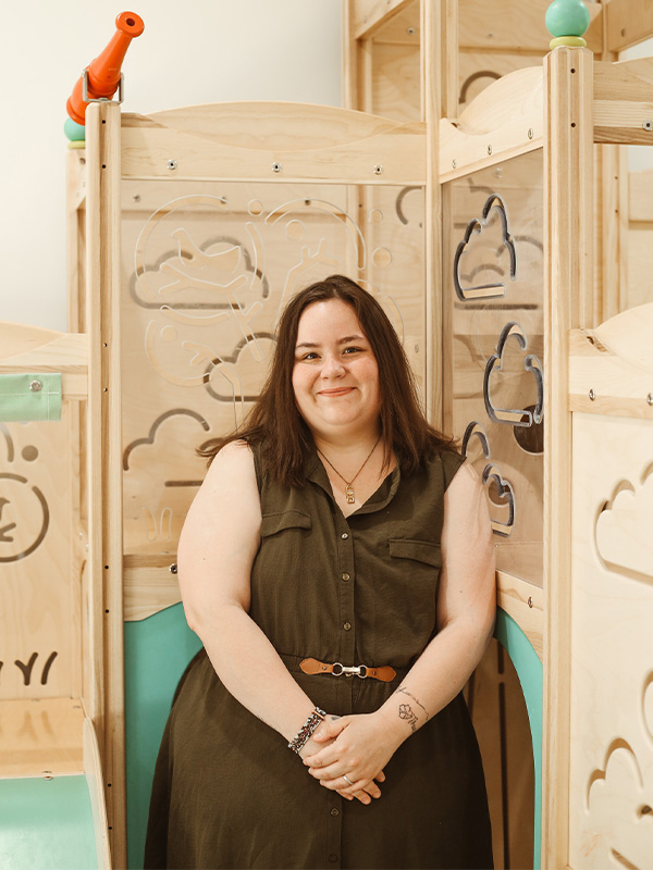 A woman with long brown hair in a sleeveless olive dress sits and smiles in a wooden play area with cloud designs and a red toy periscope in the background.