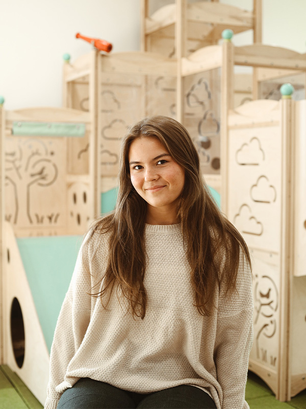 A young woman with long brown hair, wearing a beige sweater, sits and smiles in front of a wooden play structure decorated with drawings of clouds and trees. The setting appears bright and playful.