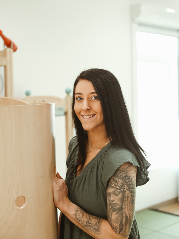 A woman with long dark hair and arm tattoos, wearing a green dress, stands indoors and smiles at the camera while holding a light wooden structure.