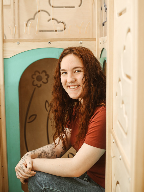 A young woman with long curly red hair, wearing a red t-shirt and jeans, sits and smiles inside a wooden play structure decorated with flower and cloud designs.