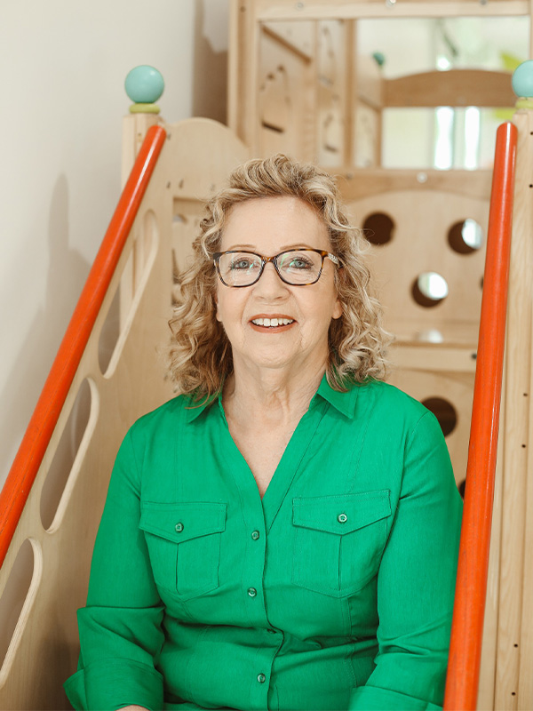 An older woman with curly blonde hair and glasses, wearing a bright green shirt, sits smiling on an indoor wooden play structure with orange rails.