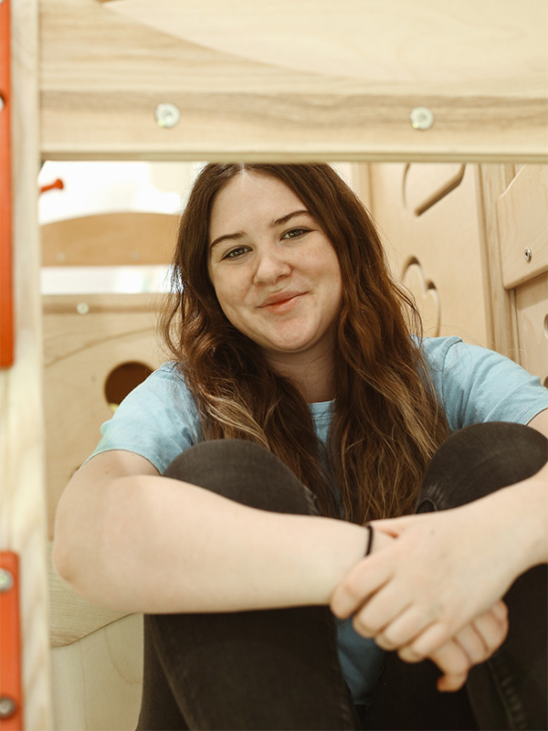 A young woman with long brown hair sits cross-legged, smiling, inside a wooden play structure. She is wearing a light blue t-shirt and black pants. The background shows light-colored wooden panels.