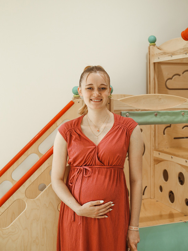 A pregnant woman in a red dress smiles while standing in front of a wooden playground structure indoors, gently resting one hand on her belly.