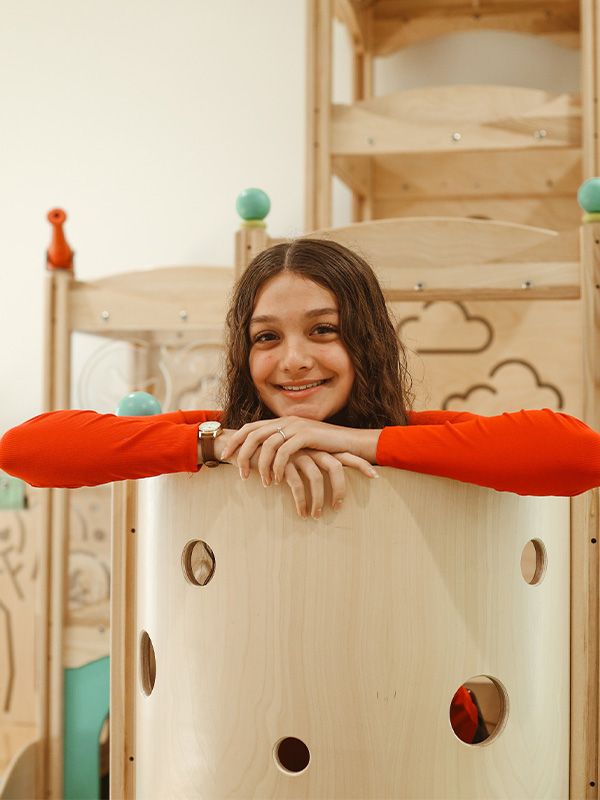 A smiling girl with curly hair and a red long-sleeve shirt leans on a wooden play structure with circular cutouts, in a bright indoor setting.