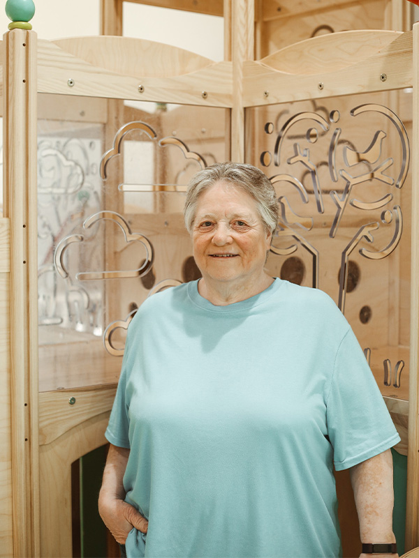 An older woman with short gray hair, wearing a light blue shirt, stands in front of a wooden panel decorated with carved tree and cloud designs, smiling at the camera.