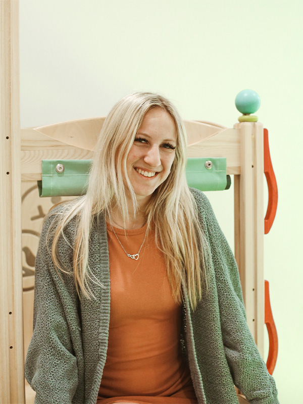 A woman with long blonde hair, wearing an orange dress and gray cardigan, sits and smiles in front of a light-colored wooden structure with colorful accents.