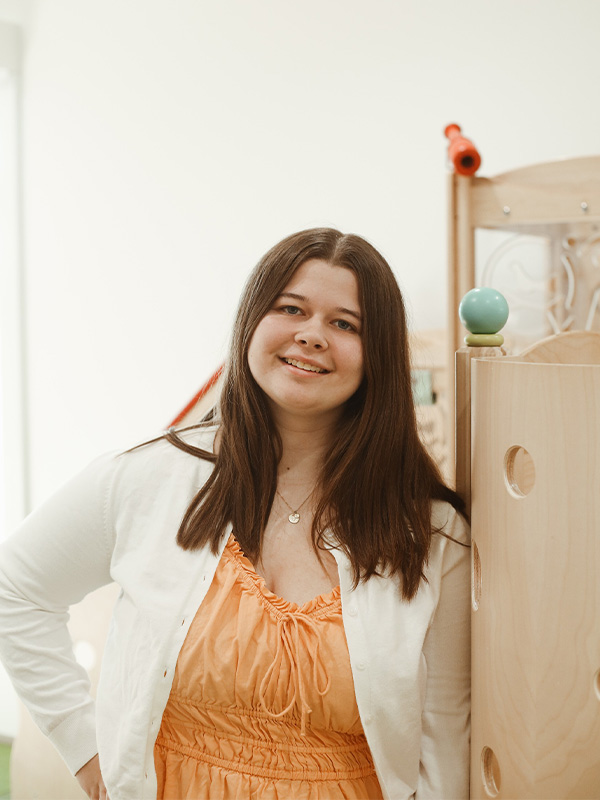 A woman with long brown hair, wearing an orange dress and white cardigan, smiles while standing indoors next to a wooden structure with a small blue ball on top.