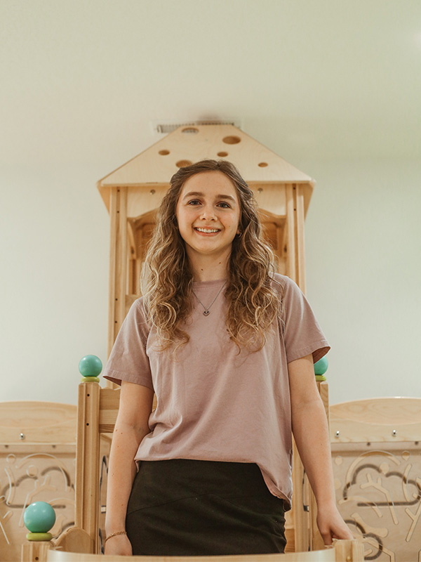 A young woman with long wavy hair, wearing a mauve t-shirt and black skirt, smiles while standing in front of a wooden play structure indoors.