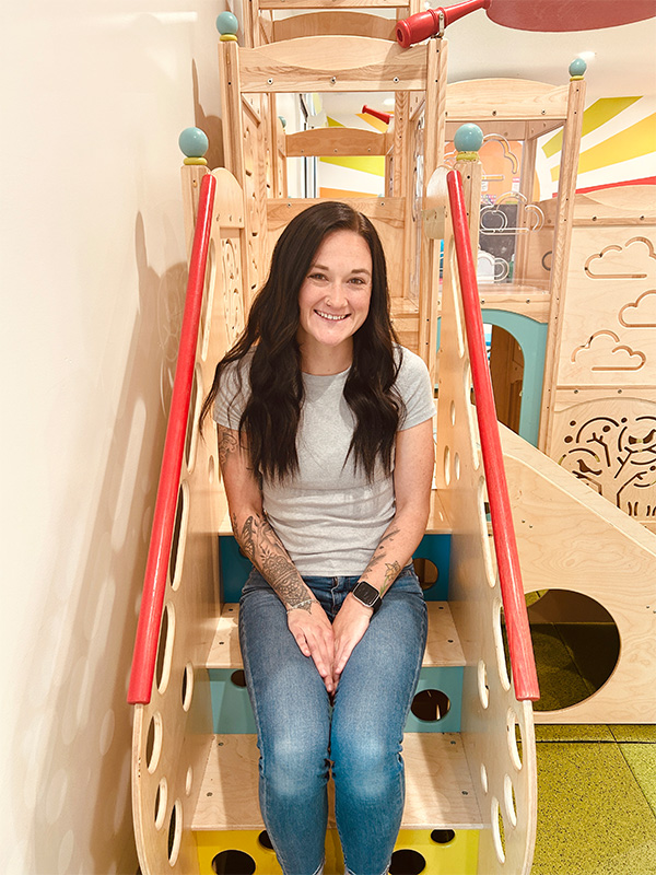 Kayla with long dark hair wearing a gray t-shirt and blue jeans sits smiling on wooden steps in a colorful indoor play area shaped like a ship.