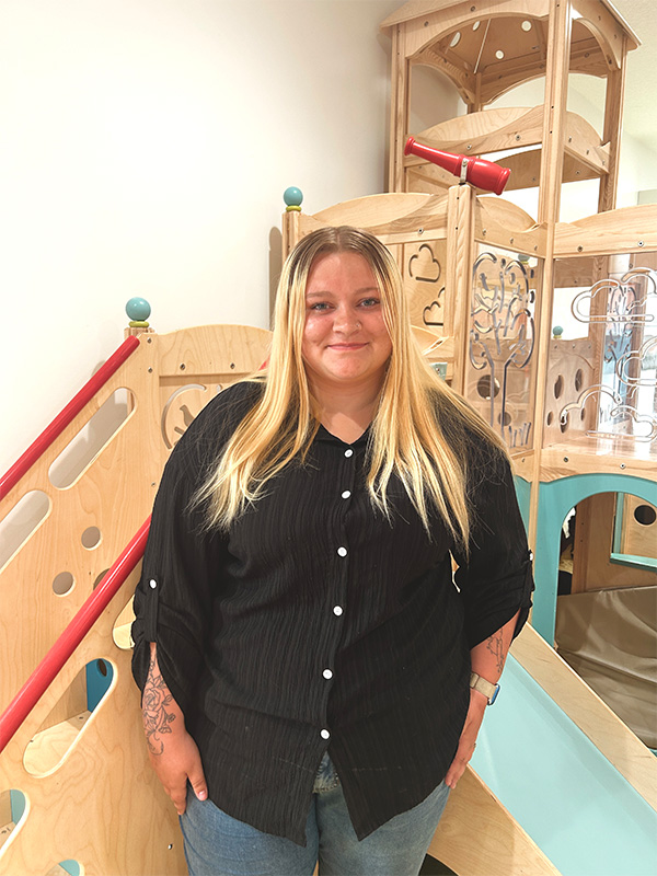 Gracey with long blonde hair, wearing a black button-up shirt and jeans, stands smiling in front of a light wood indoor playground structure.