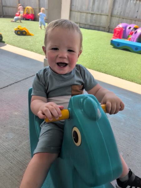 A smiling toddler in a blue outfit rides a teal rocking toy outdoors. Other children and colorful toys are visible in the background on a playground with artificial grass.