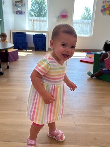 A smiling toddler in a colorful striped dress and pink sandals stands on a wooden floor in a bright, sunny classroom with large windows and children playing in the background.