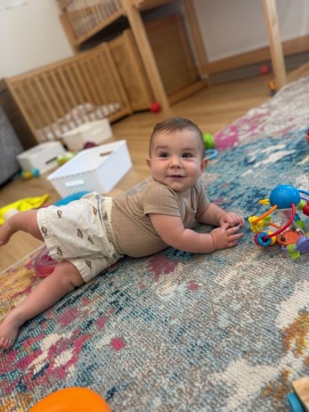 A baby in light-colored shorts and a tan shirt lies on a colorful rug, smiling and propped on one arm, surrounded by toys in a cozy play area with a crib and basket in the background.