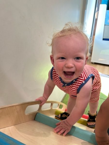 A smiling toddler with blonde hair in a red, white, and blue striped outfit climbs a small indoor wooden ramp, looking up excitedly at the camera.