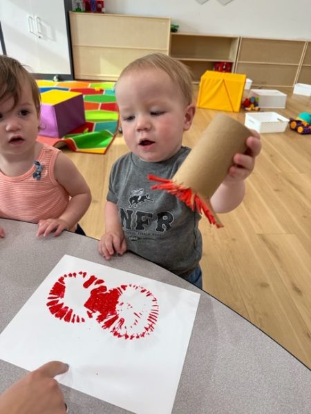 A toddler holds a cardboard tube with red paint on the end, stamping it on white paper to make prints. Another young child watches, and colorful toys are visible in the background.