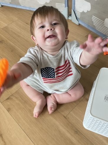 A smiling baby sits on a wooden floor wearing a shirt with an American flag, reaching forward with both hands. A white basket and a playpen with cloud patterns are nearby.