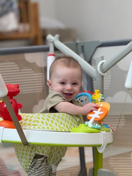 A smiling baby sits in a colorful activity jumper, surrounded by toys, including a red animal and an orange toy with a white cross shape. The background is softly blurred, showing a crib and other baby items.