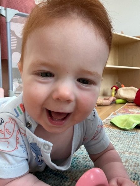 A smiling baby with light skin and red hair wearing a light-colored onesie with animal prints is lying on their stomach on a patterned rug, with toys and shelves visible in the background.