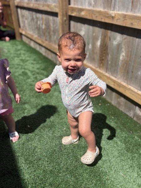 A toddler in a patterned romper and Crocs stands on artificial grass, smiling and holding a small toy. A wooden fence and another child's partial figure are visible in the background.