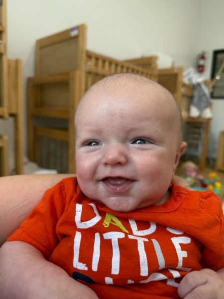 A smiling baby wearing an orange shirt with white and yellow letters is held up indoors. Wooden cribs and toys are visible in the softly lit background.