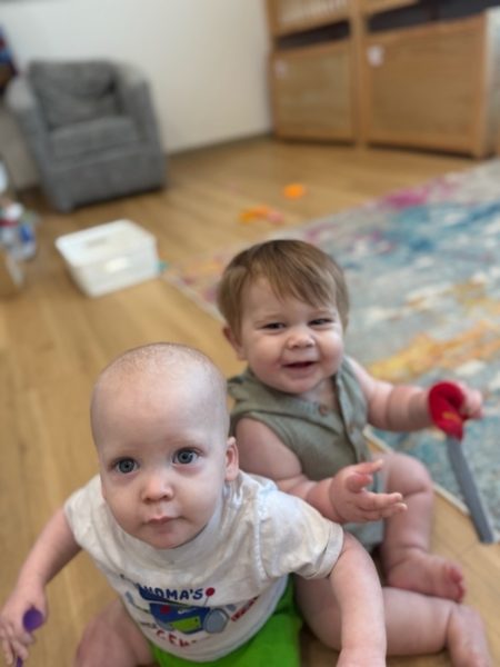 Two babies sit on a colorful rug indoors. The baby in front wears a white shirt and looks at the camera, while the baby behind, smiling, wears a sleeveless outfit and holds a toy.