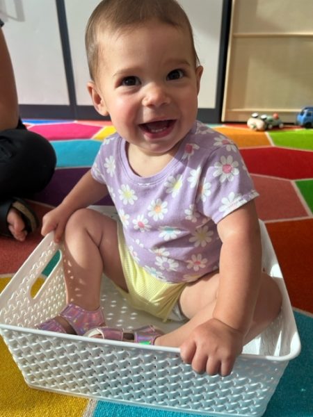 Smiling baby in a purple floral shirt and yellow shorts sits inside a white plastic basket on a colorful tiled carpet, with toys and shelves in the background.