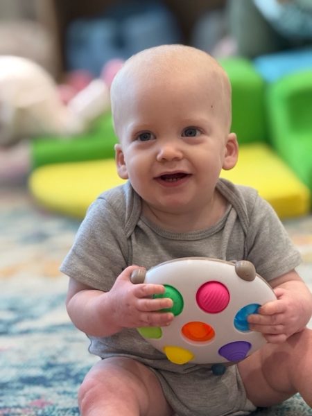 A smiling baby in a gray onesie sits on the floor, holding a colorful toy with large, round buttons. The background features soft, out-of-focus play mats and toys.