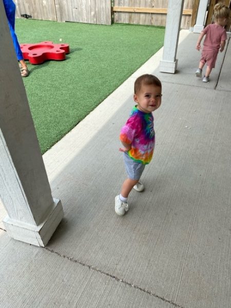 A toddler in a colorful tie-dye shirt and gray shorts smiles while walking on a concrete patio near artificial grass. Another child is visible in the background, also walking away.