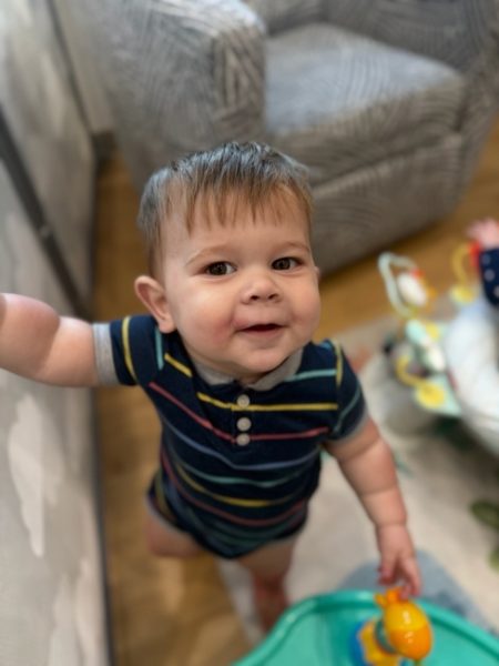 A smiling baby with short brown hair and a striped onesie stands indoors, holding a yellow toy. A patterned armchair and baby play area are visible in the background.