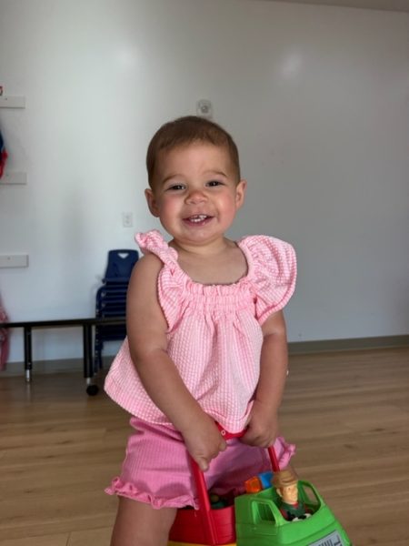 A smiling toddler wearing a pink outfit stands on a wooden floor, holding a green and red toy truck. The background shows a small table, stacked chairs, and a plain wall.