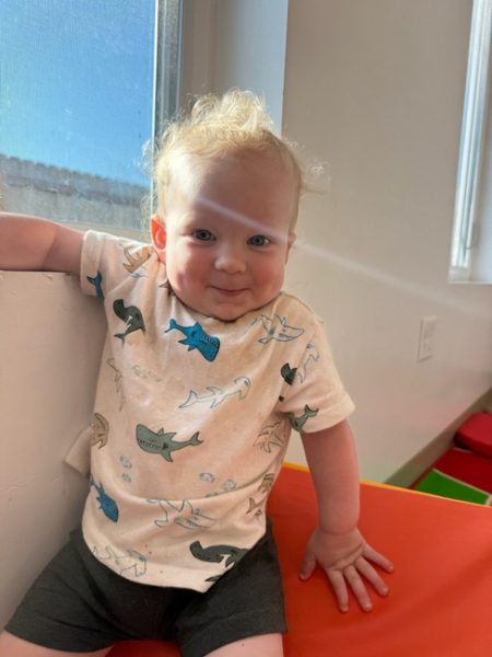 A smiling toddler with curly blond hair sits on a red and orange mat by a sunlit window, wearing a shirt decorated with cartoon sharks and dark shorts.