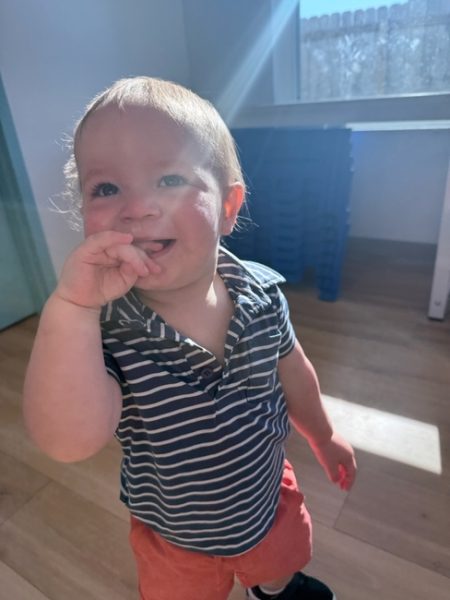 A smiling toddler in a striped shirt and red shorts stands indoors, with sunlight streaming in and casting shadows on the wooden floor. The child is holding their hand near their mouth and looking up.