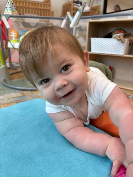 A smiling baby with light brown hair lies on a blue mat, wearing a white shirt. The baby looks up at the camera, with toys and shelves visible in the background of a playroom.