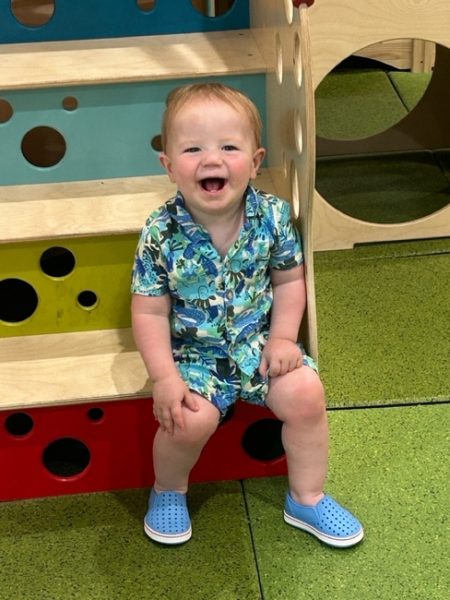 A smiling toddler wearing a blue floral shirt and matching blue shoes sits on colorful, perforated wooden steps in a play area with green flooring.