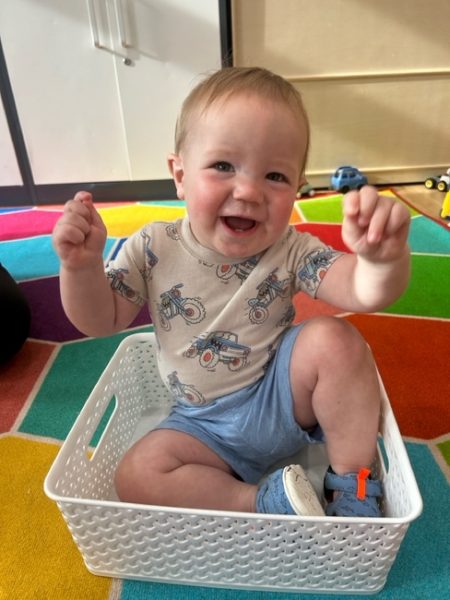 A smiling baby wearing a shirt with motorcycles and blue shorts sits inside a white plastic basket on a colorful, patterned rug. The baby has arms raised and looks happy and playful.