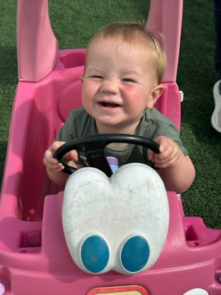 A smiling toddler sits in a pink toy car, holding the steering wheel with both hands. The car has large white and blue features on the front, and the child appears happy and playful.
