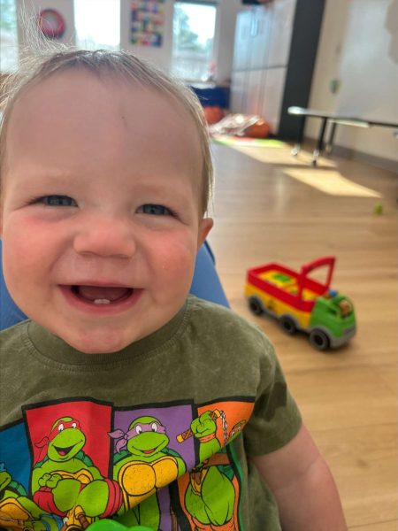 A smiling baby wearing a Teenage Mutant Ninja Turtles t-shirt sits indoors on a wooden floor, with a colorful toy truck nearby and sunlight streaming through windows in the background.