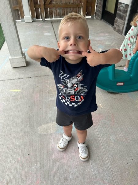 Young boy standing on a patio, pulling the sides of his mouth with his fingers to make a silly face. He wears a navy shirt with race cars, black shorts, and white shoes. A girl and playground equipment are in the background.