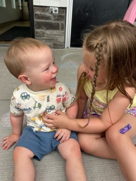 A young boy and girl sit on a concrete surface, smiling at each other. The boy wears a vehicle-patterned shirt and blue shorts, while the girl wears a yellow dress with braided hair and a purple bandage on her knee.