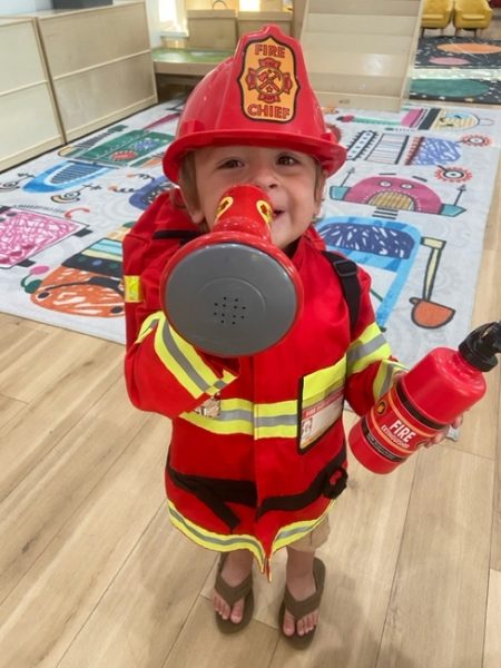 A young child dressed as a firefighter, wearing a red fire chief helmet and coat, smiles while holding a toy megaphone and fire extinguisher in a colorful, playful indoor setting.