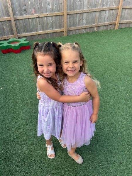 Two young girls with long hair, wearing light purple dresses, stand on green artificial grass and hug each other, smiling at the camera. A wooden fence and play equipment are visible in the background.