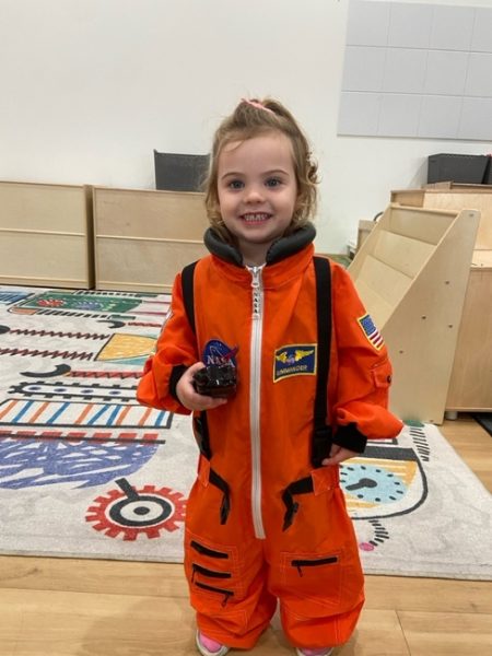 A young girl wearing an orange astronaut suit with NASA patches smiles while standing indoors on a colorful rug with geometric patterns. She is holding a small toy and appears to be in a playroom.