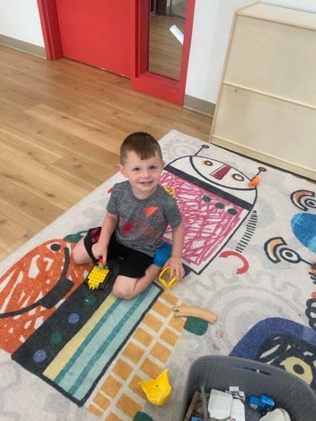 A young boy sits on a colorful rug with playful designs, smiling while holding a toy construction vehicle. Nearby are scattered toys and a basket of blocks, with a red door and wooden shelving in the background.