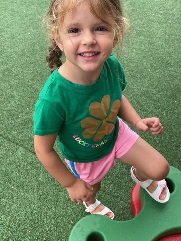 A young girl with light brown hair in a ponytail, wearing a green "Lucky Charms" shirt, pink shorts, and white sandals, smiles while standing on playground equipment with artificial grass beneath her.