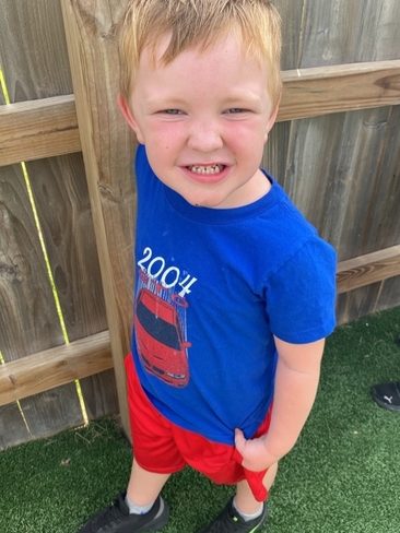 Young boy with blond hair wearing a blue "2004" shirt with a red car graphic, red shorts, and black sneakers stands smiling on green turf near a wooden fence.