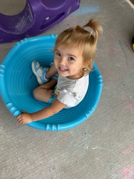 A smiling young child with light hair sits inside a blue plastic tub on a concrete surface, looking up at the camera. Colorful chalk drawings and playground equipment are visible around her.