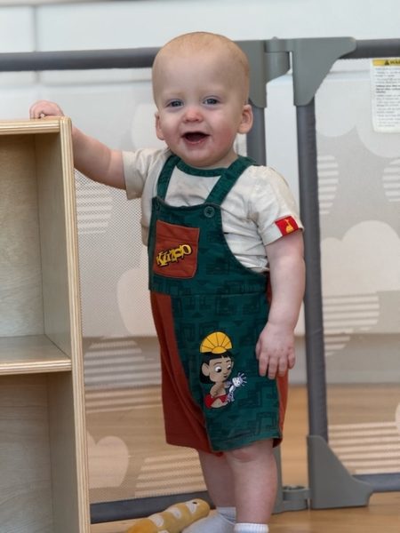 A smiling baby stands next to a wooden shelf, wearing green and orange overalls with a cartoon character on them. A gray baby gate is visible in the background.