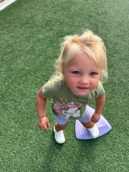 A young child with blonde hair, wearing a green unicorn t-shirt, light shorts, and white shoes, stands on green artificial grass next to a purple stepping stone, looking up and smiling.