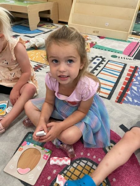 A young girl in a pastel-colored dress sits on a colorful classroom rug, holding puzzle pieces and looking up at the camera. Other children and puzzle pieces are visible around her.