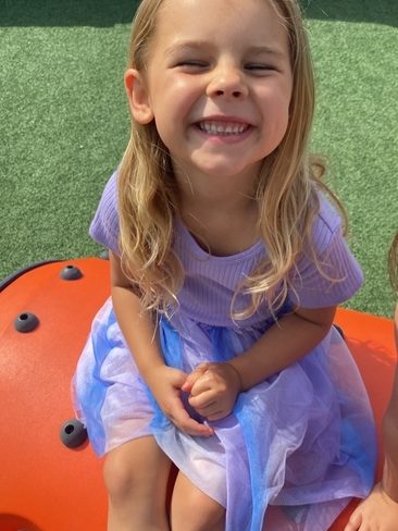 A young girl with long blonde hair in a purple top and blue tulle skirt smiles widely while sitting on an orange playground structure, with green artificial grass and a wooden fence in the background.